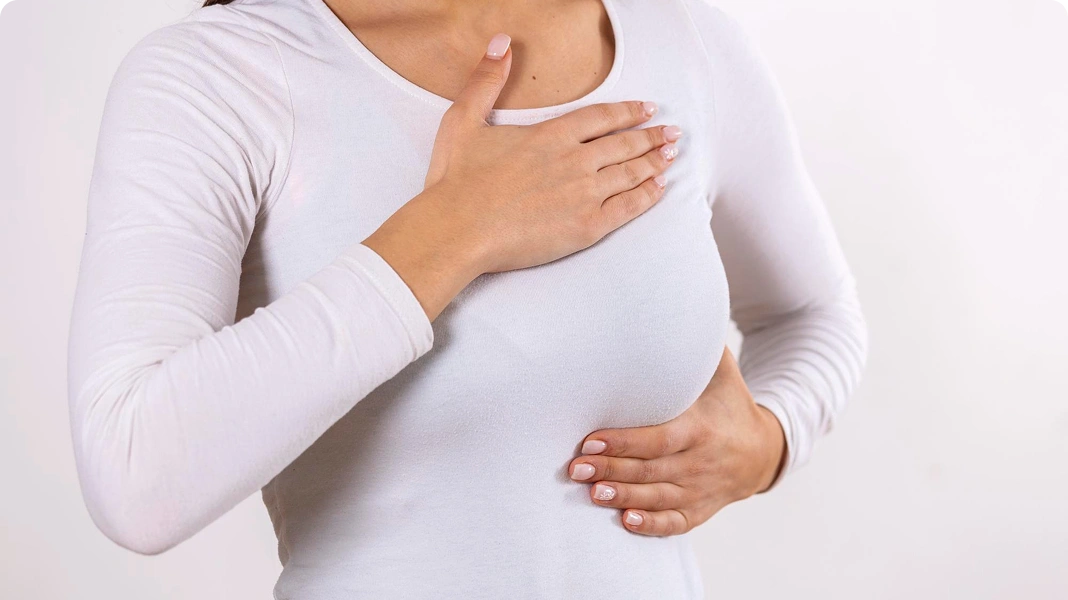 Woman wearing a white t-shirt performing a self-breast exam by feeling her left breast with both hands.