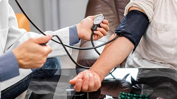 A hand holds a sphygmomanometer gauge while another person’s arm is wrapped in a blood pressure cuff, measuring blood pressure.