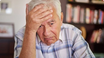 An older man touching his forehead and looking confused or distressed, with a healthcare professional offering comfort